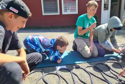 vier Jungs knien auf dem Boden und bauen eine Trage im Survivalcamp